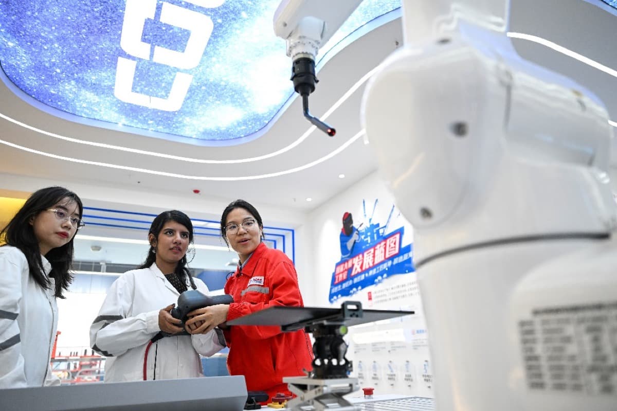 Inside an industry exhibition hall, three visitors get a hands on look at a white industrial robotic arm as a staff member explains, with a screen reading development blueprint behind them.
