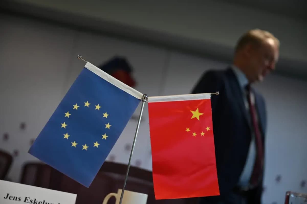 一场会议桌上摆放的欧盟与中国迷你旗帜特写,背景中一名男子的身影若隐若现。