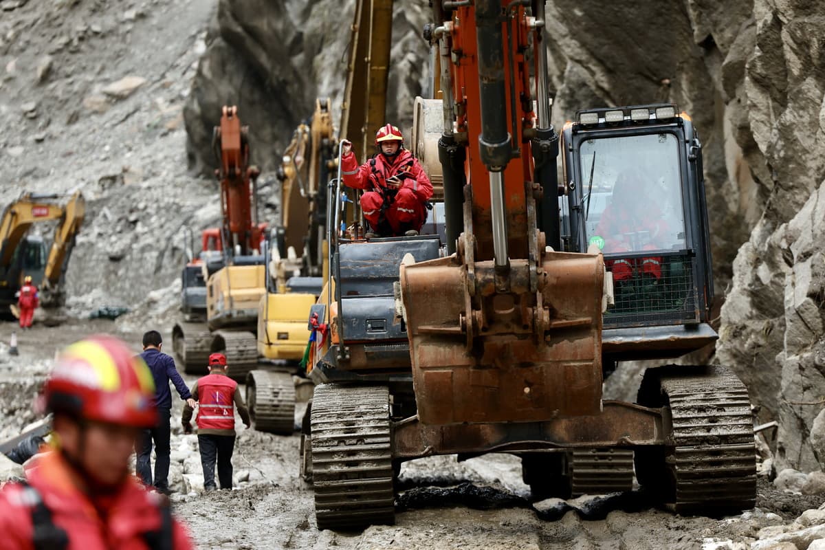 Along a road hemmed in by steep cliffs, a line of excavators works in tandem to clear accumulated mud and rock, as rescue personnel direct the operation nearby.