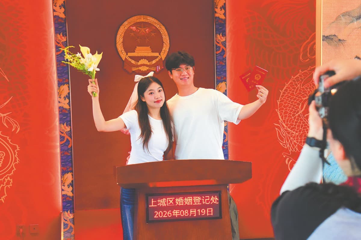 A newlywed couple holds up flowers and their marriage certificates at a registration office in Hangzhou's Shangcheng district, as a photographer captures the moment behind them.