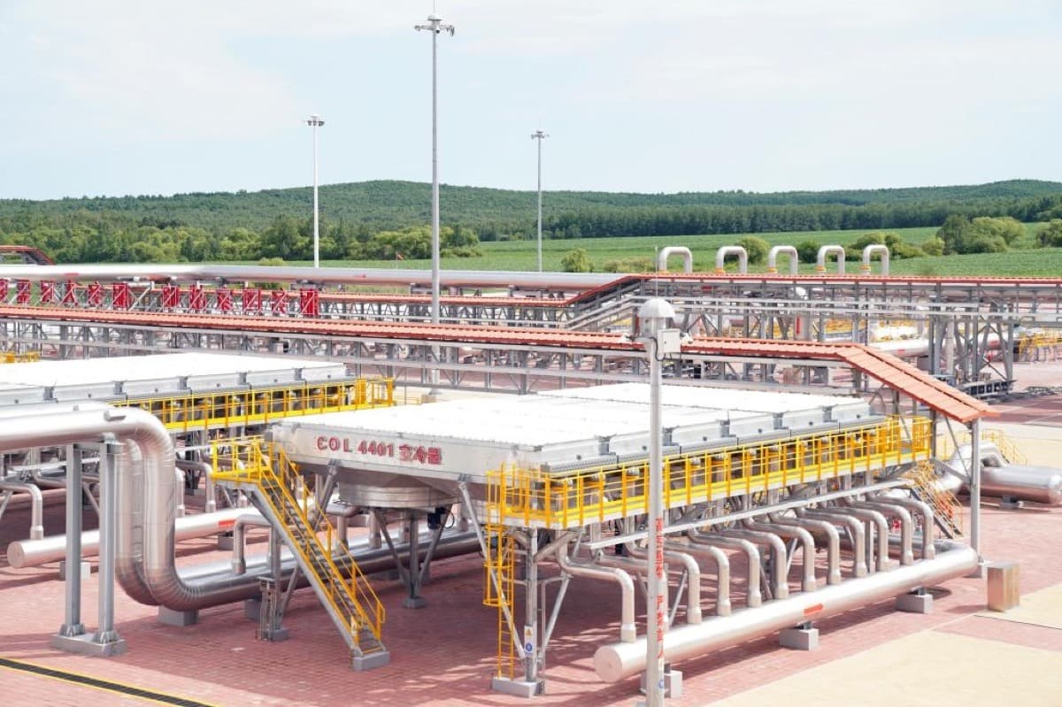 At a natural gas processing facility, rows of silver pipelines and yellow railings stretch under the sun, with green hills and plant infrastructure visible in the distance.