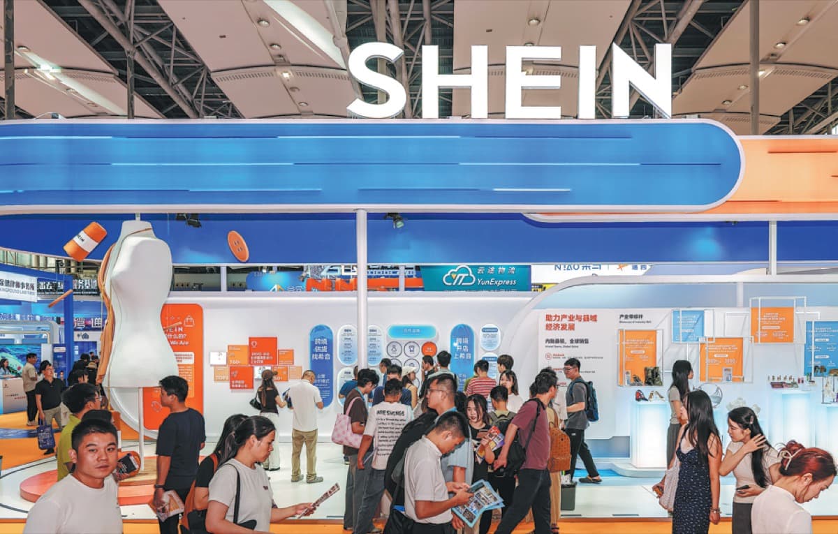 Shein's booth at an expo in Guangzhou, with a large white SHEIN sign mounted above the stand and crowds of visitors browsing below.